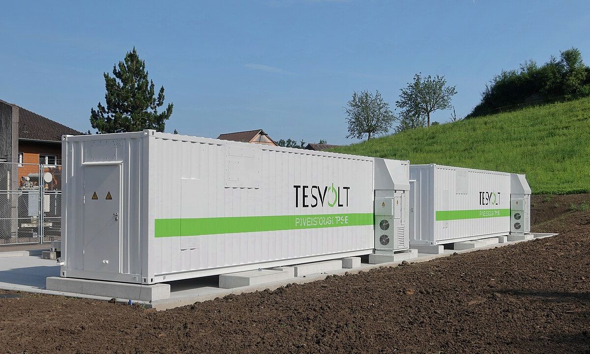 Kontenerowe magazyny energii stojące na fundamencie obok budynków